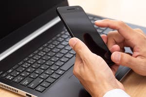 手に持ったスマートフォンを操作しながら、開いたノートパソコンのキーボードに手を置く人。