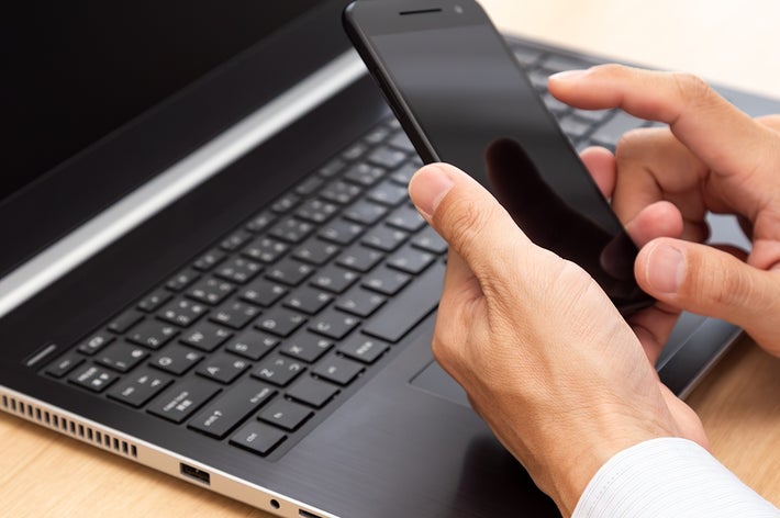 手に持ったスマートフォンを操作しながら、開いたノートパソコンのキーボードに手を置く人。