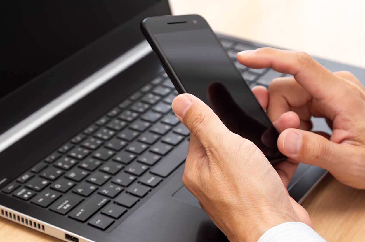 手に持ったスマートフォンを操作しながら、開いたノートパソコンのキーボードに手を置く人。