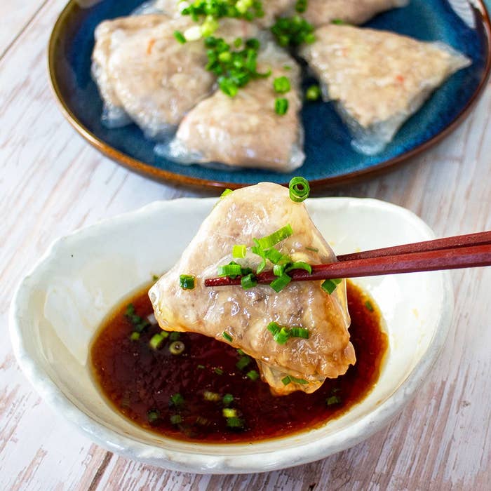 箸でつかんだ三角形の餃子がタレに入れられる瞬間。皿にもいくつか餃子が盛られている。上にネギが添えられている。