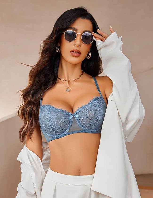 Person wearing sunglasses, bluish lace lingerie, and a achromatic jacket, posing with a assured look and long, styled hair