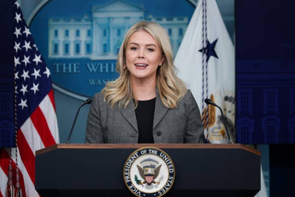 Person at podium in press briefing room, wearing a professional gray blazer, smiling, with the White House emblem in the background