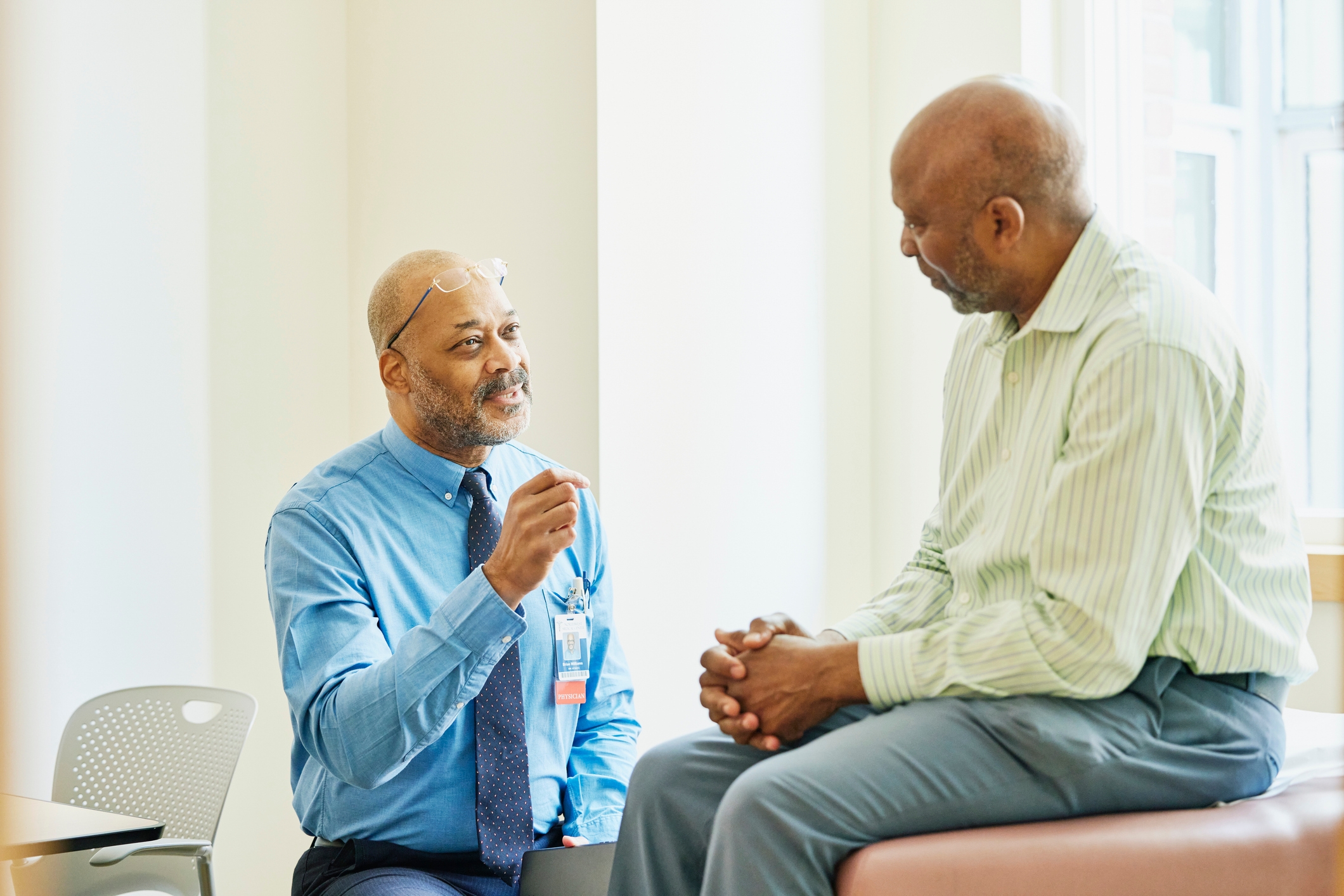 En lege i en blå skjorte og slips snakker med en eldre mann som sitter på et eksamensbord i et legekontorinnstilling