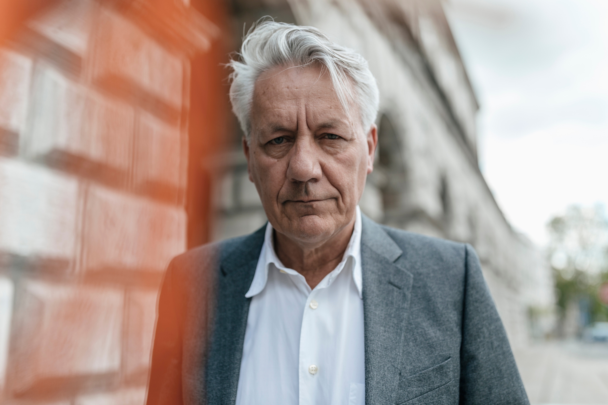 En eldre mann i dress og hvit skjorte står utendørs og ser direkte på kameraet. Bakgrunnen viser en uskarp bygning