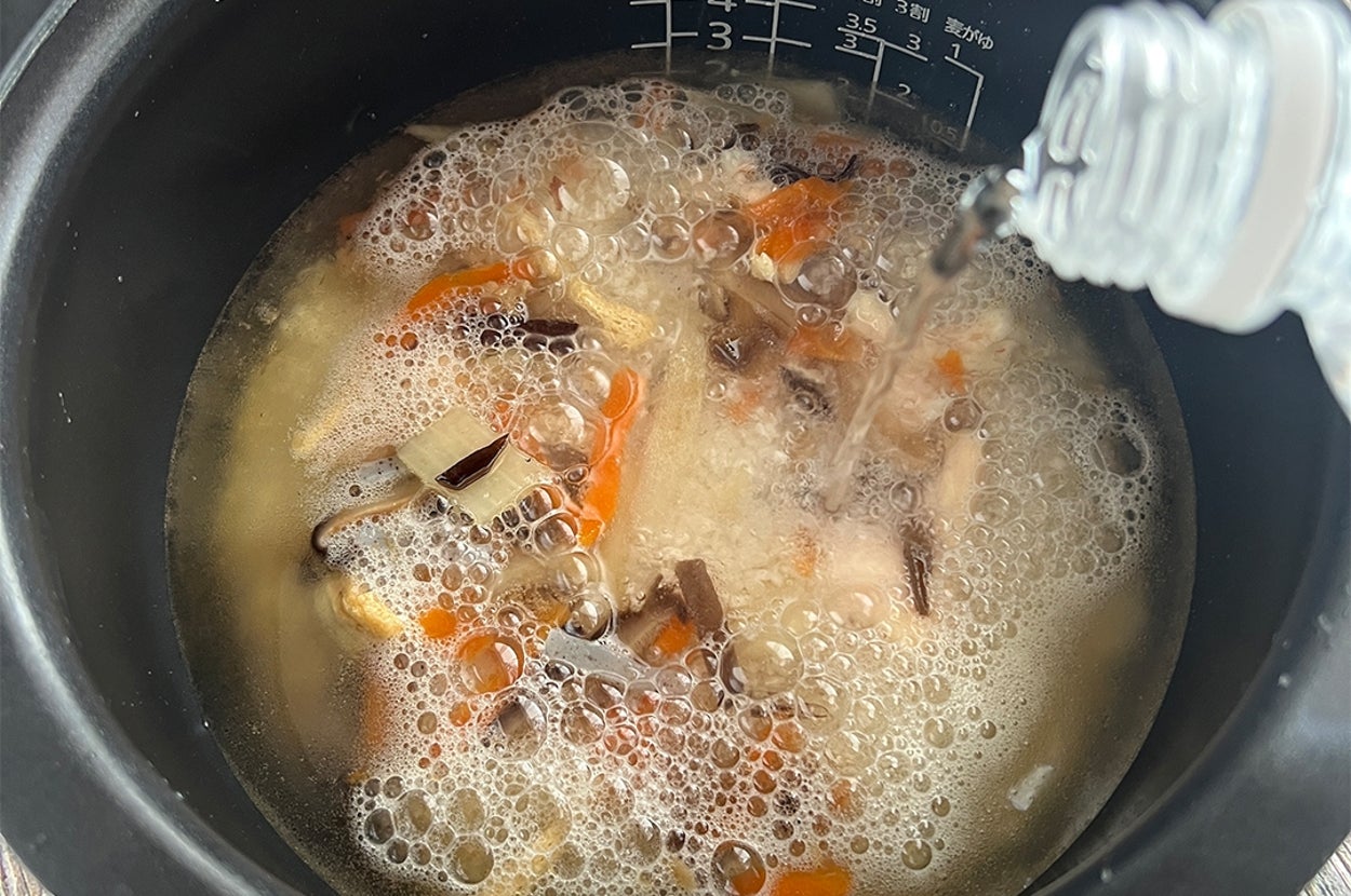 炊飯器で炊き込みご飯の準備中。野菜やキノコが見える中、液体が注がれている。