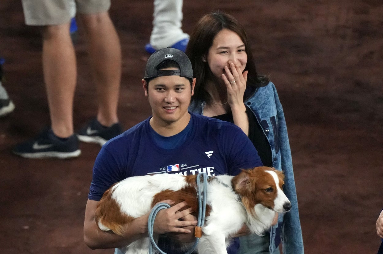 野球選手がシャツとスポーツパンツを着て、犬を抱えて微笑んでいる。後ろに少人数の観客がいる。