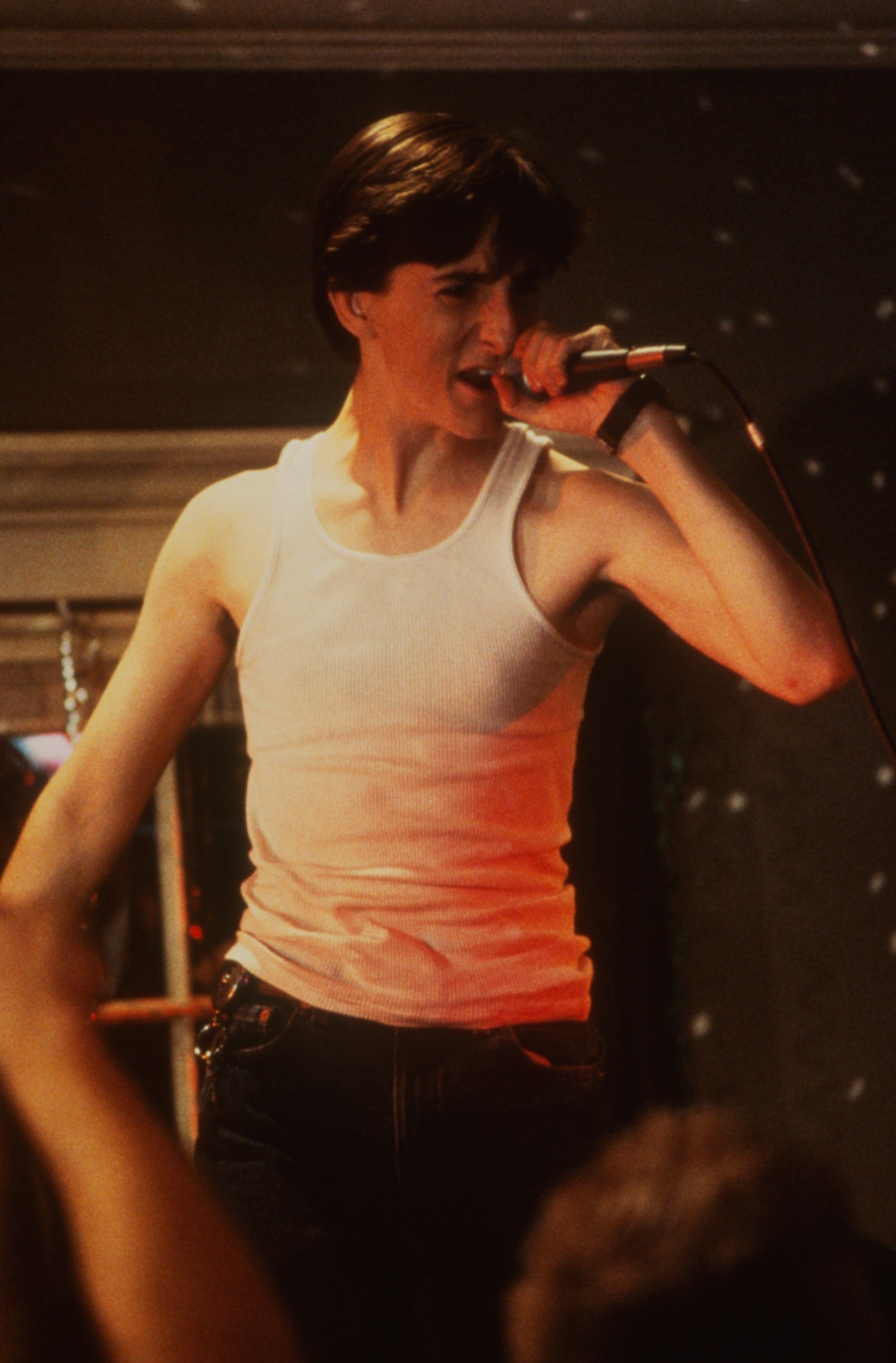 Person in a white tank top passionately singing into a microphone on stage