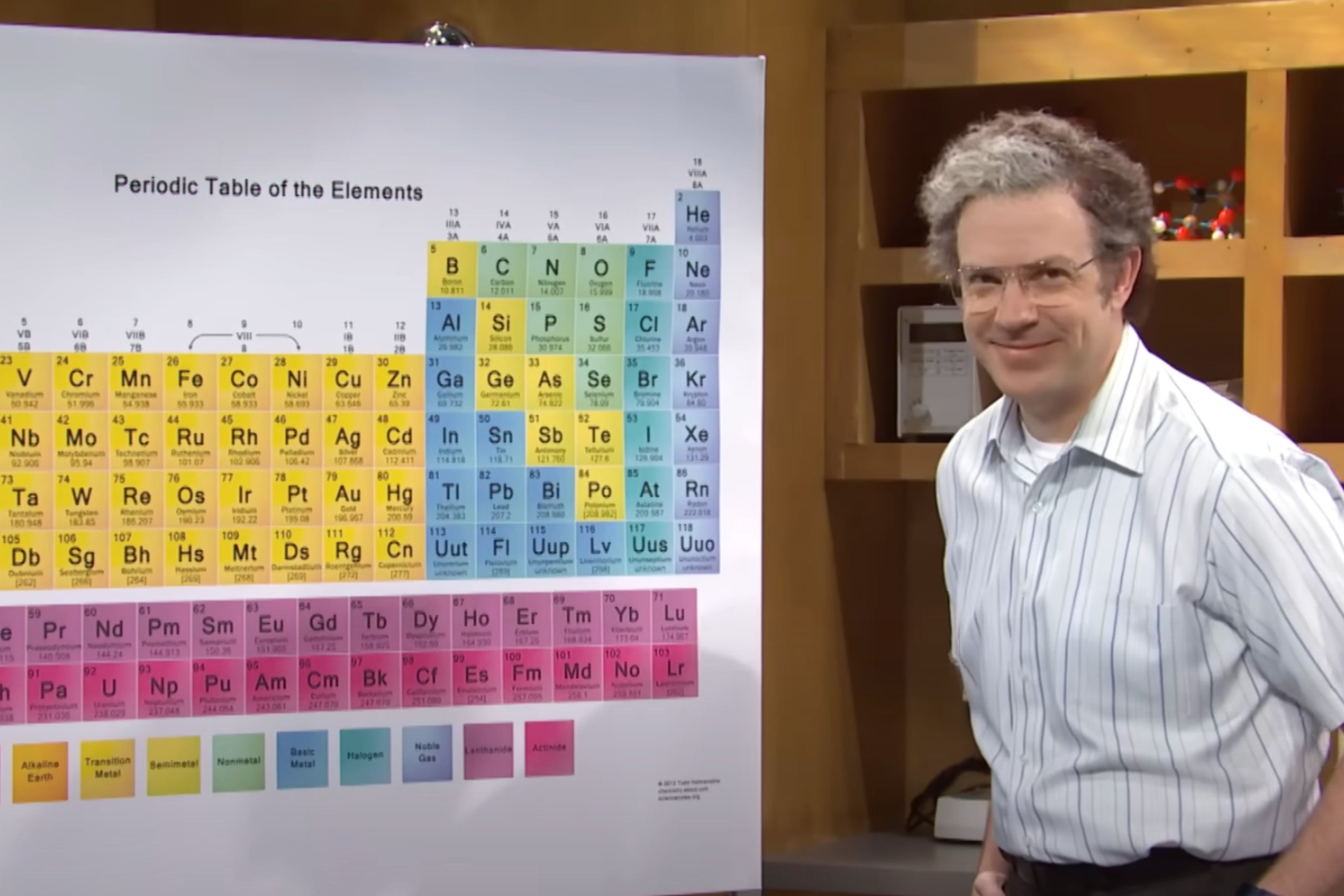 Jason Sudeikis stands beside a large periodic table poster, smiling, in an SNL sketch