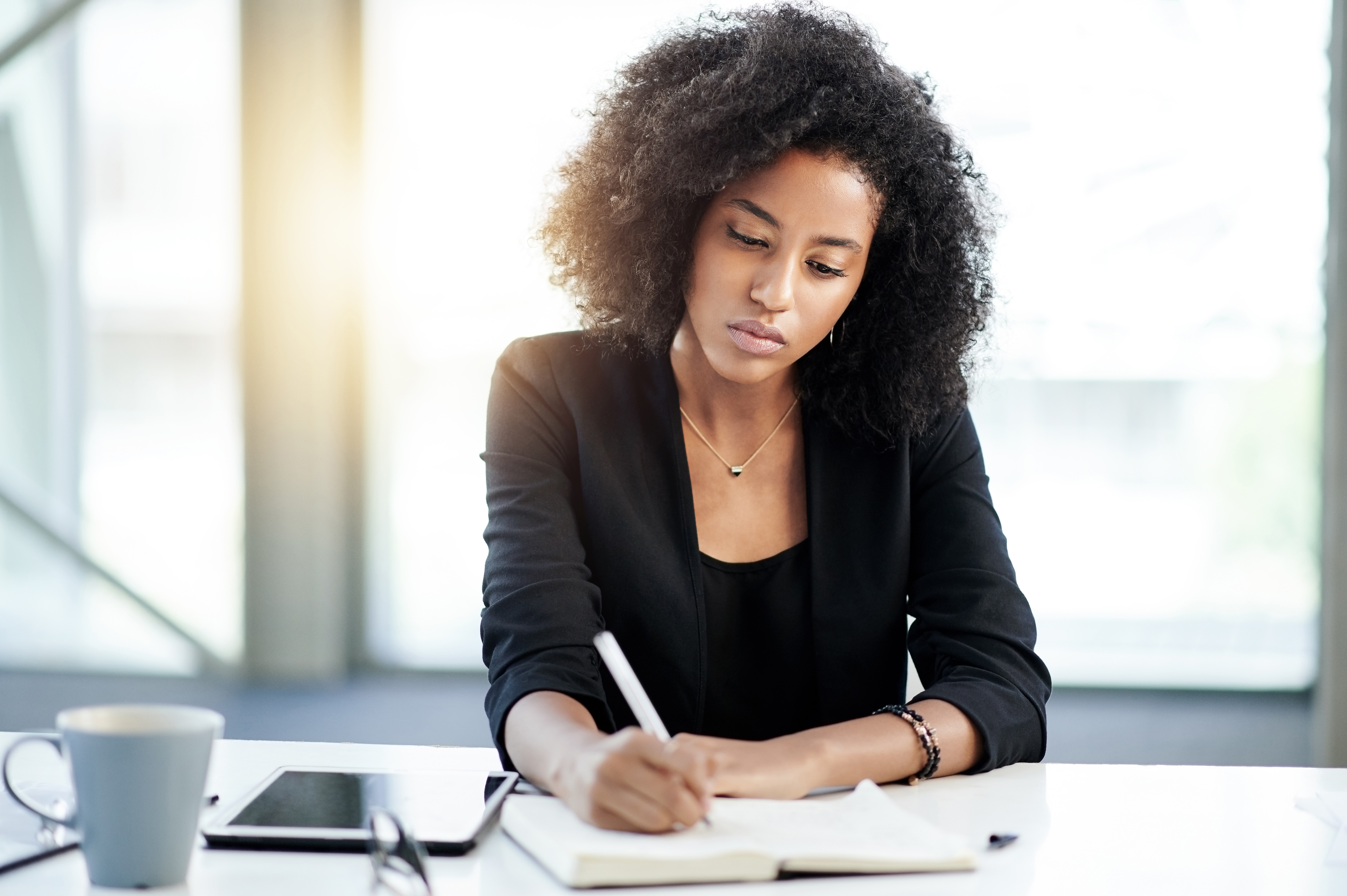 Person som sitter ved et bord, fokusert på å skrive i en notisbok, med et nettbrett og kopp i nærheten, i et rolig miljø som antyder produktivitet
