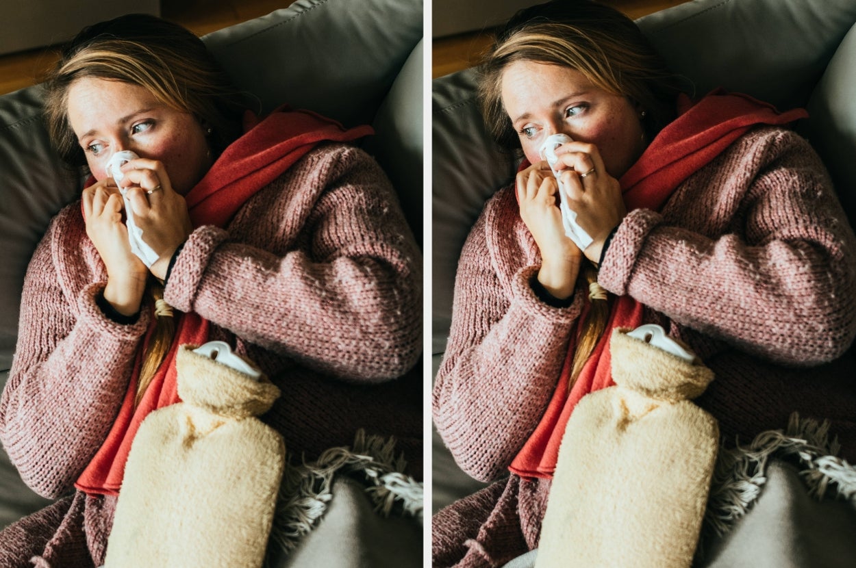 Person lying on a couch, holding a tissue to their nose, wearing a cozy sweater and red scarf, with a hot water bottle nearby