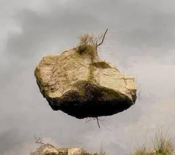 Large rock with grass appears to float mid-air, creating an optical illusion against a cloudy sky. Ground vegetation partially visible below