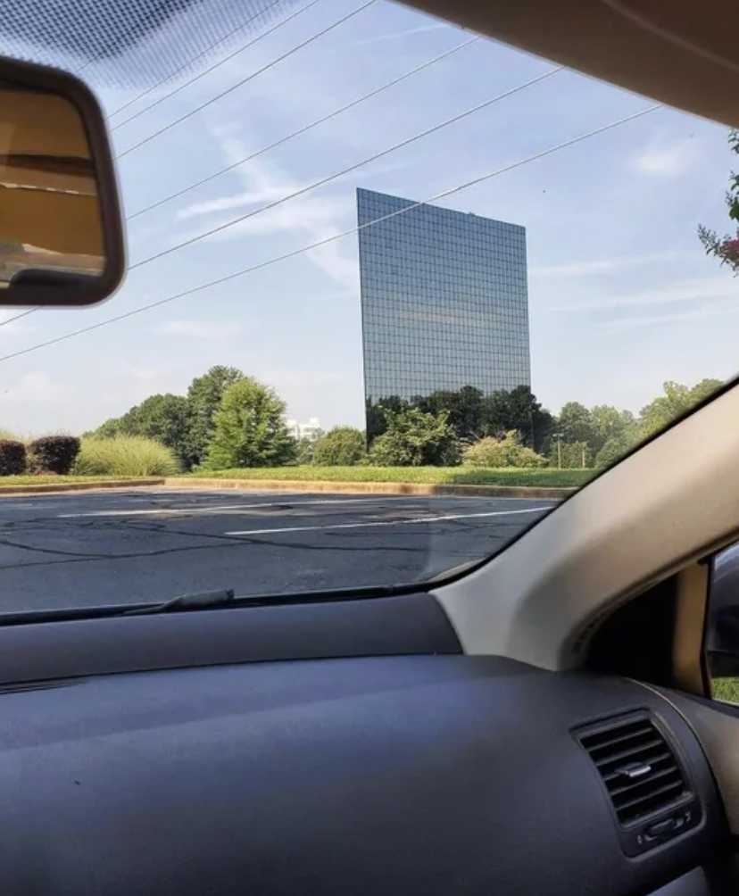 View from inside a car of a large mirrored office building reflecting the sky and trees, with trees and bushes surrounding its base