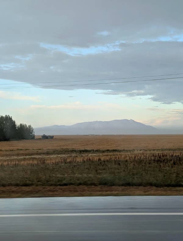 Open field with distant mountains under a cloudy sky, viewed from a roadside