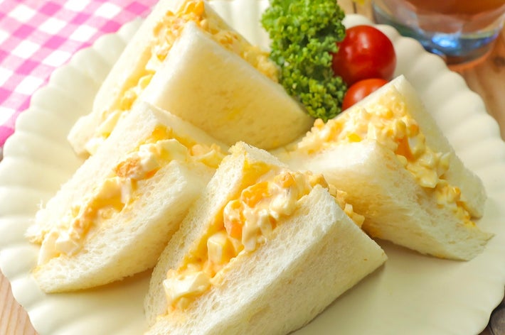 卵サラダサンドイッチが白い皿に盛られ、パセリとミニトマトが添えられています。背景には飲み物があります。