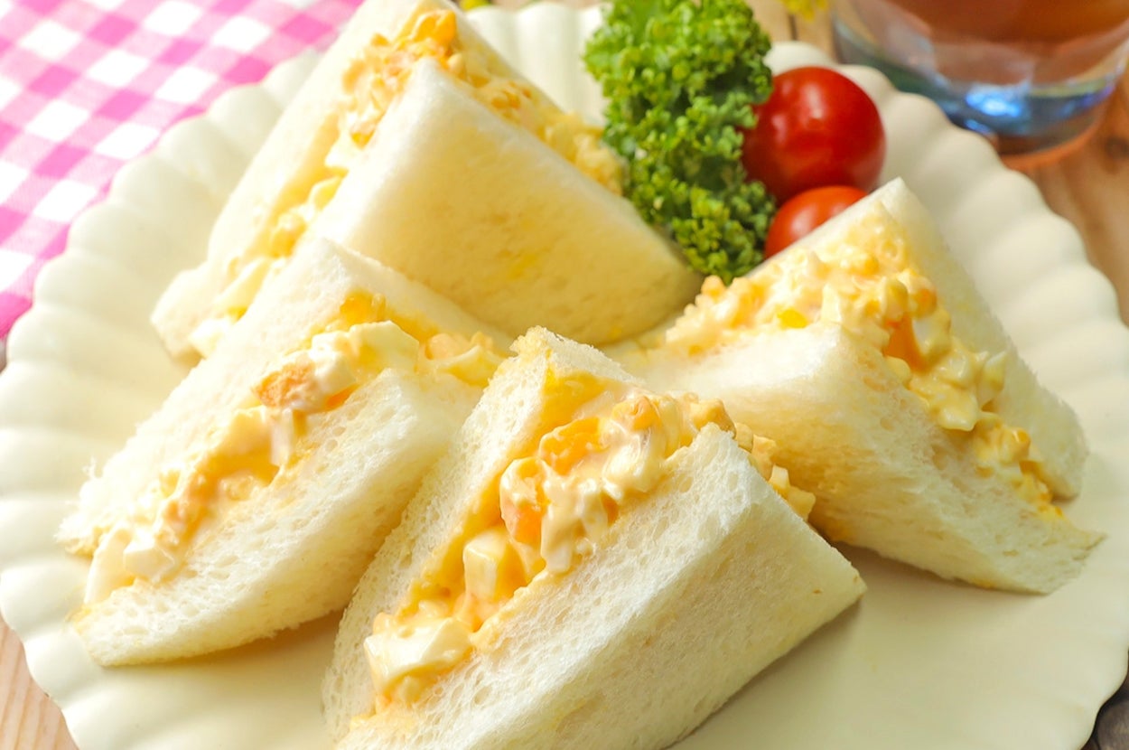 卵サラダサンドイッチが白い皿に盛られ、パセリとミニトマトが添えられています。背景には飲み物があります。