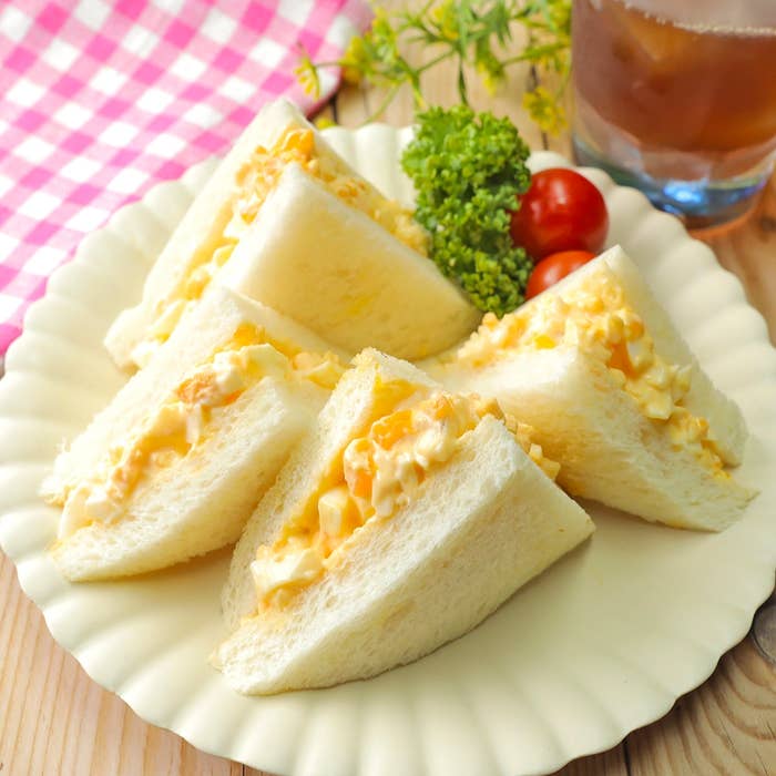 卵サラダサンドイッチが白い皿に盛られ、パセリとミニトマトが添えられています。背景には飲み物があります。