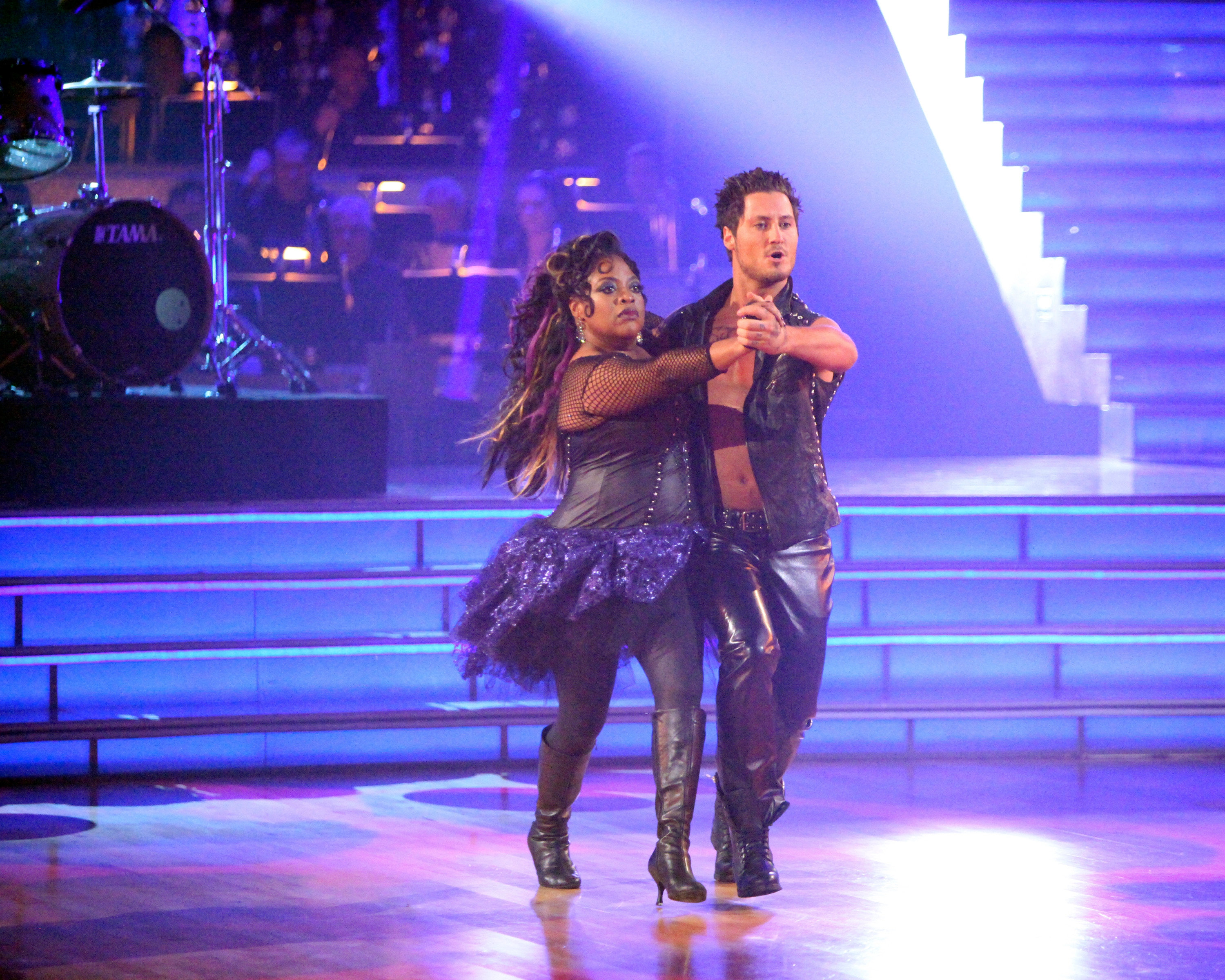 Two dancers perform on a lit stage, wearing coordinated, edgy leather outfits with embellishments