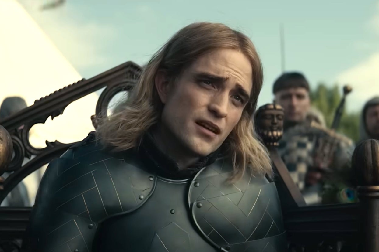 Robert Pattinson in "The King" in medieval armor with long hair, appearing thoughtful in an outdoor scene with others blurred in the background