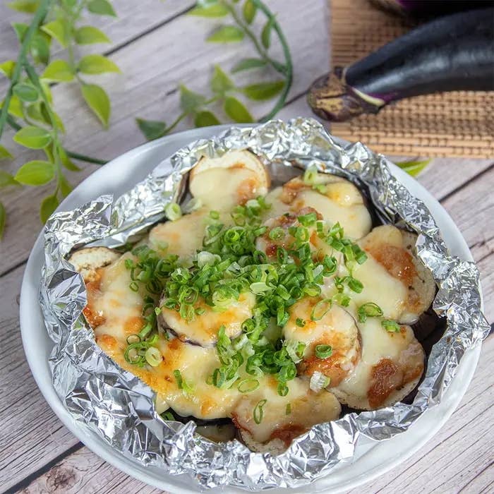 アルミホイルに包まれた焼きなすの上に溶けたチーズと刻んだねぎがのっている料理。