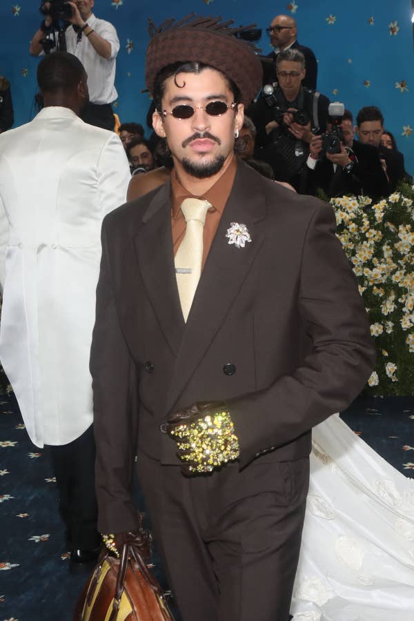 Bad Bunny wearing a dark suit with gold embellishments, sunglasses, and a hat on a stylish event carpet. Surrounded by photographers