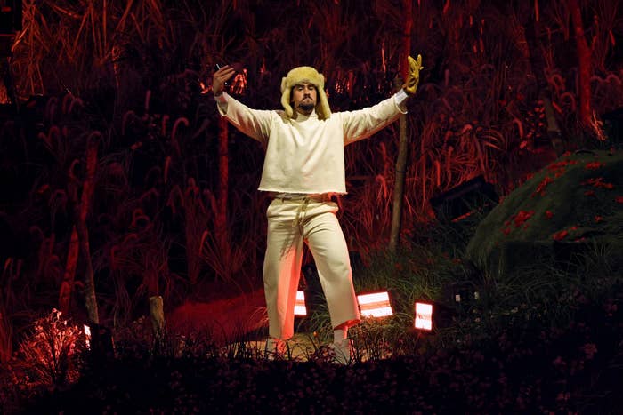 Bad Bunny on stage in winter hat and layered outfit, arms raised dramatically amidst a nature-themed setting with lighting accents
