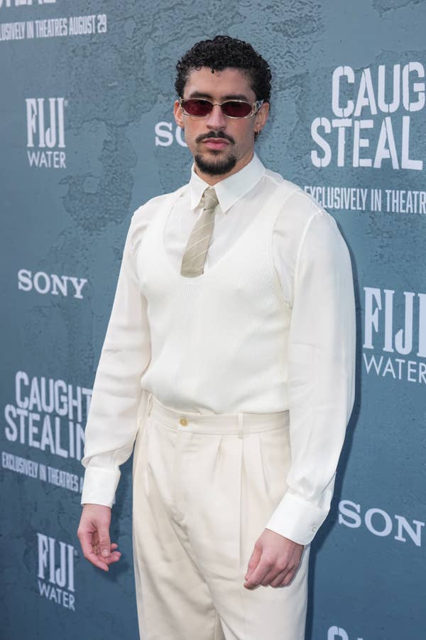 Bad Bunny in formal attire with a sleeveless white sweater and tie, posing in front of a backdrop with film logos and text