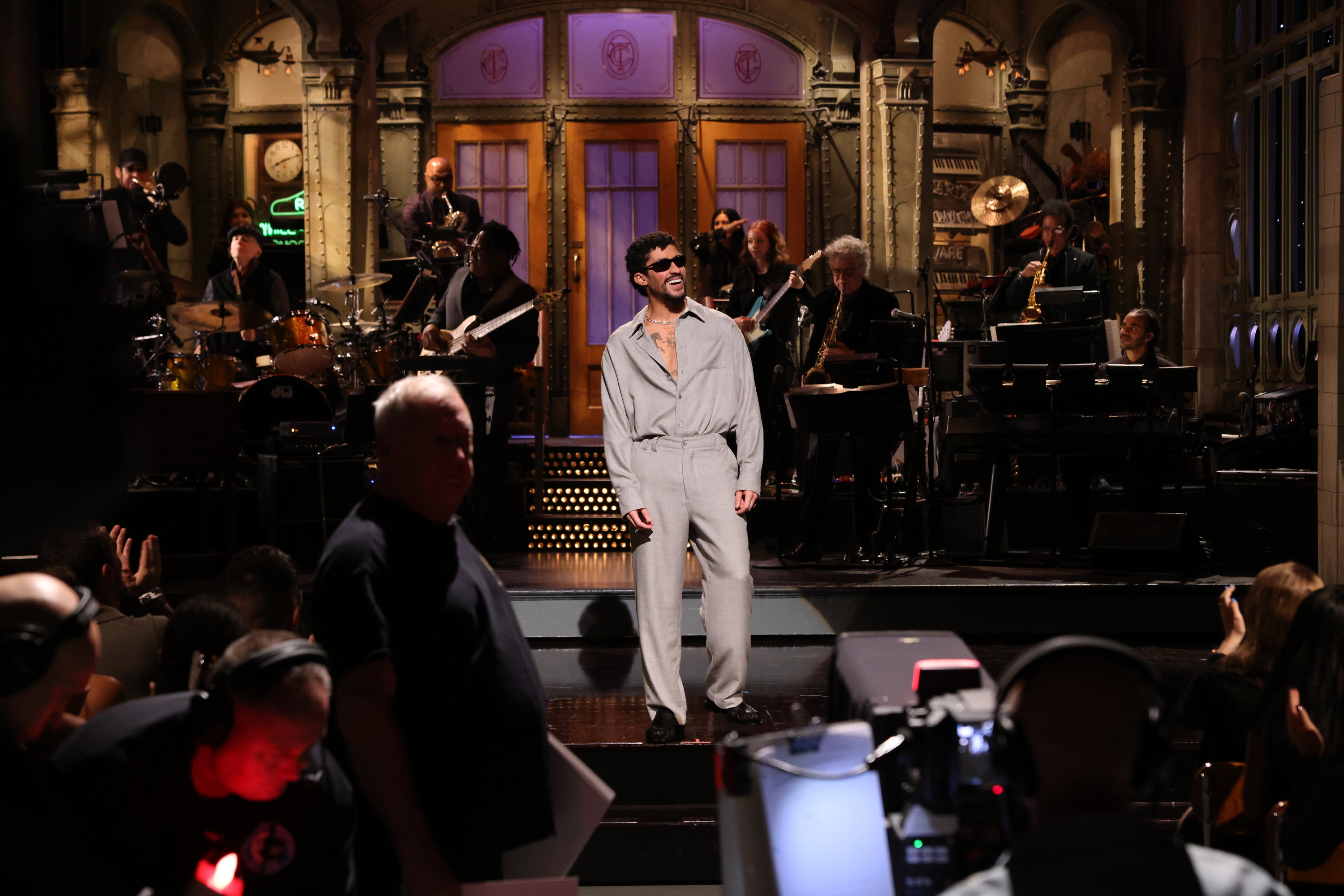 Bad Bunny on stage performing with a live band on a late-night TV set. Audience and crew present