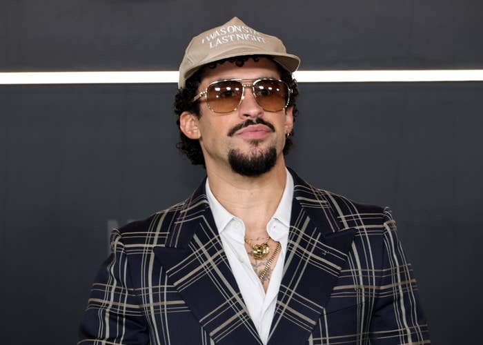 Bad Bunny wearing a plaid blazer, white shirt, and cap with sunglasses on, standing in front of a plain background