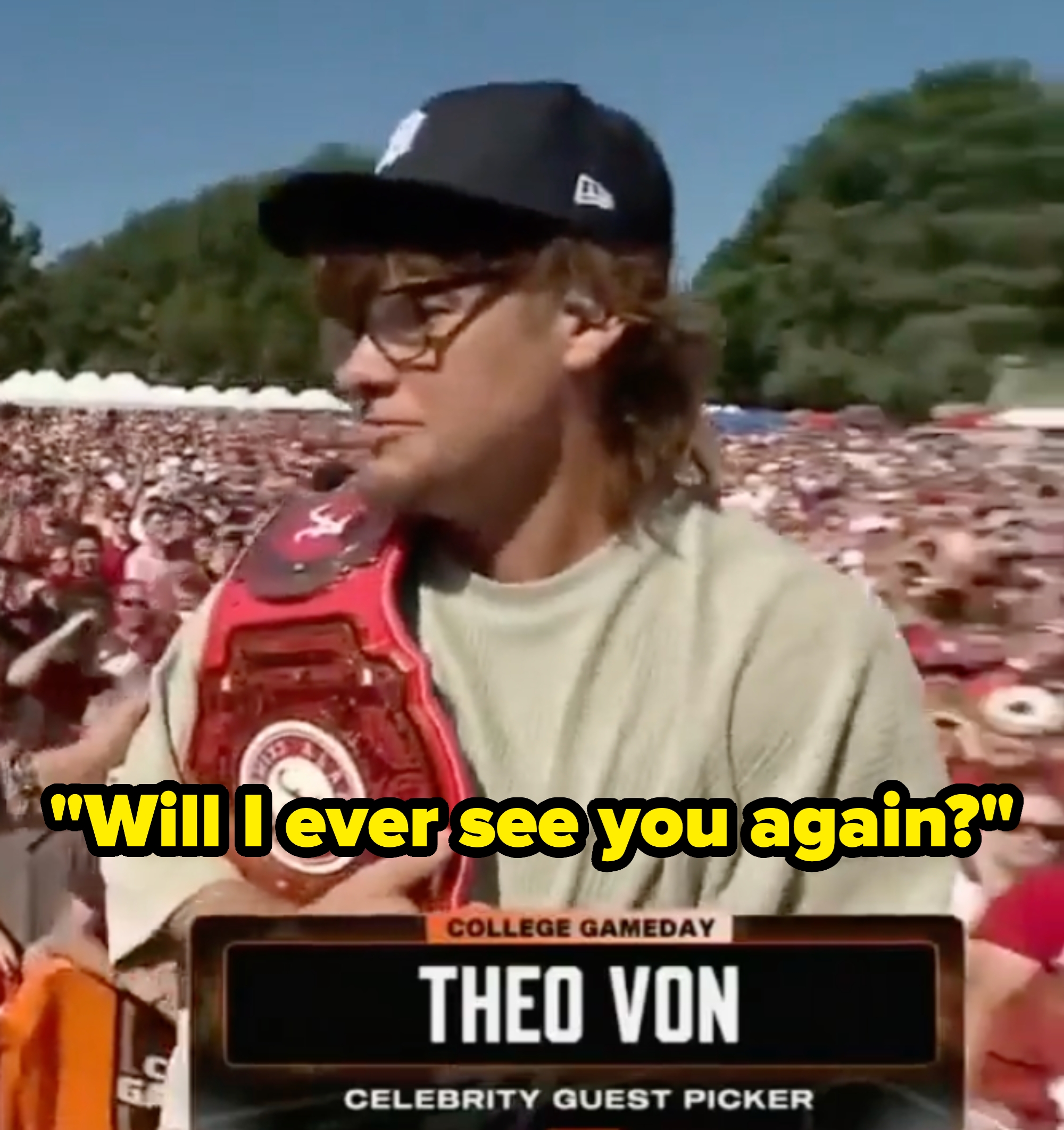 Theo Von, wearing casual attire and a cap, holds a large championship belt at a crowded outdoor event with a "College Gameday" banner