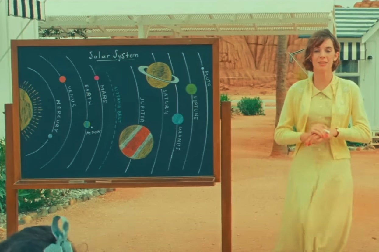 Maya Hawke in "Asteroid City" stands next to a chalkboard displaying a solar system diagram outdoors. She wears a light cardigan over a dress
