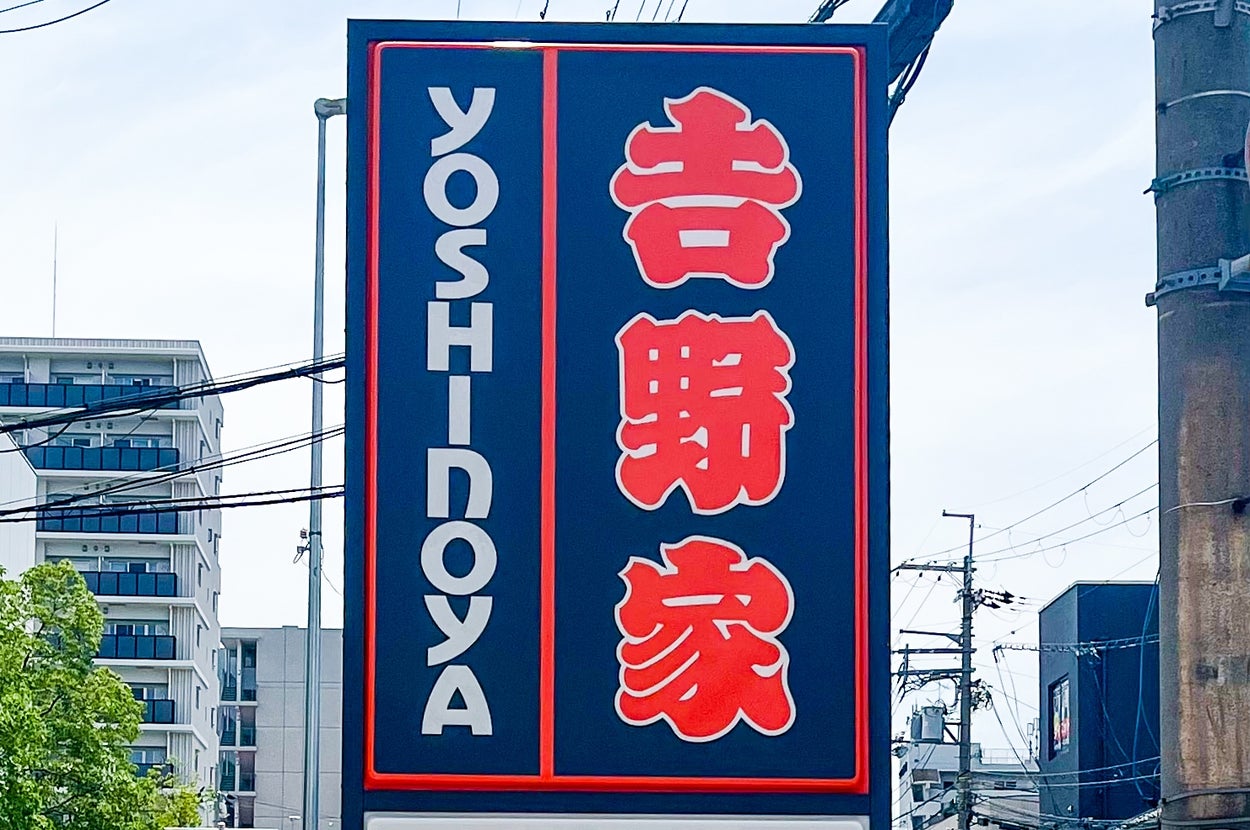 看板に「吉野家」と「YOSHINOYA」と書かれています。背景には建物と電線が見えます。