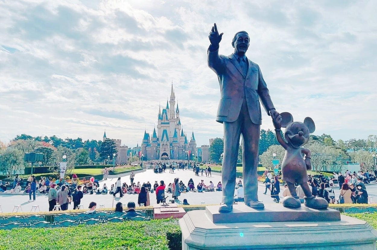 ディズニーランドでウォルト・ディズニーとミッキーマウスの像が手をつなぎ、遠くにシンデレラ城が見える賑やかな景色。