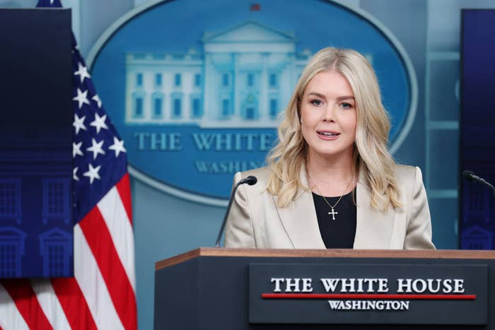 Person speaking at a podium with a White House backdrop and U.S. flag in the background. Wearing a light-colored blazer and a cross necklace