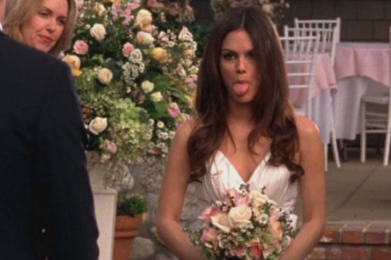 A woman in a white wedding dress playfully sticks out her tongue while holding a bouquet of flowers, with floral decorations in the background