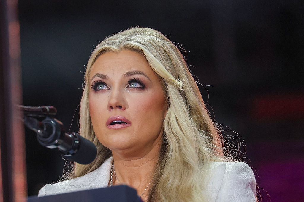 Person med langt, bølget hår, snakker ved en mikrofon, ser oppover, iført en lysfarget blazer