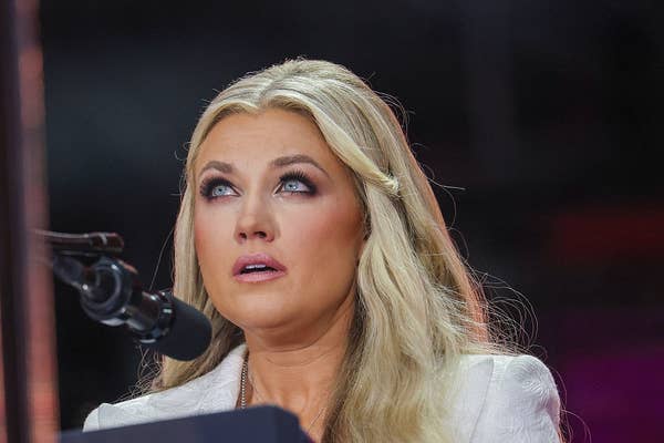 Person with long, wavy hair, speaking at a microphone, looking upwards, wearing a light-colored blazer