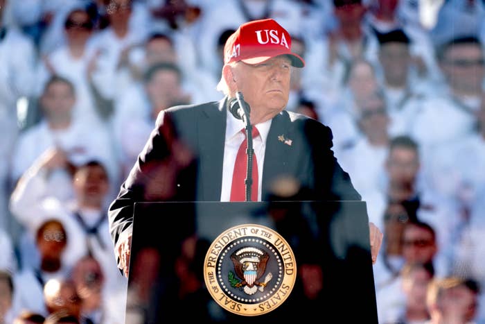 Person speaking at a podium with the presidential seal, wearing a suit and a USA cap, in front of an audience in uniform