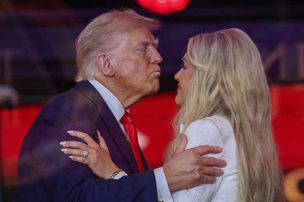 Two people embrace and lean in as if to kiss at an event. One person is wearing a suit, and the other is in a patterned outfit