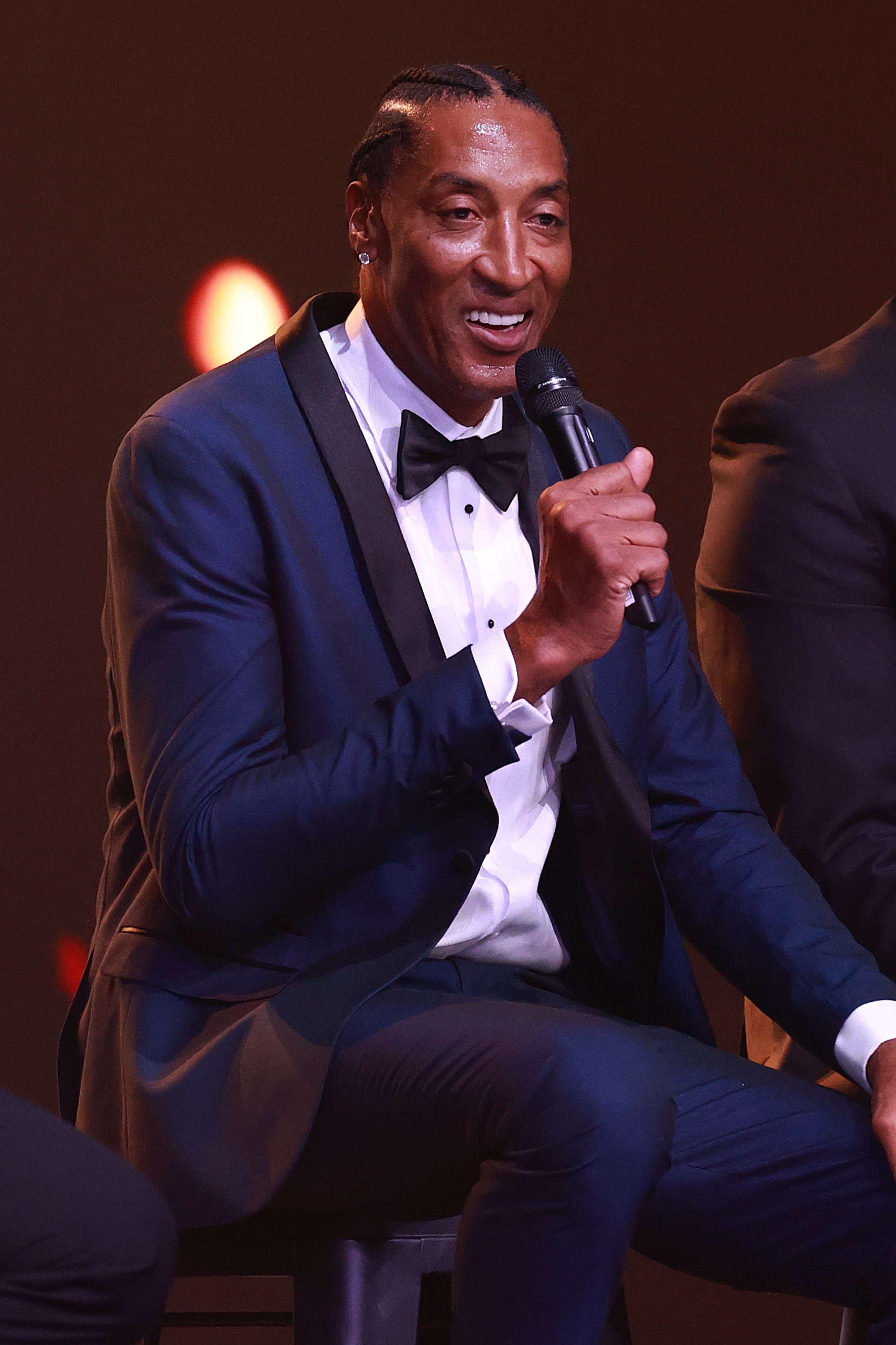 Person successful  a tuxedo sits, holding a microphone, speaking astatine  an event
