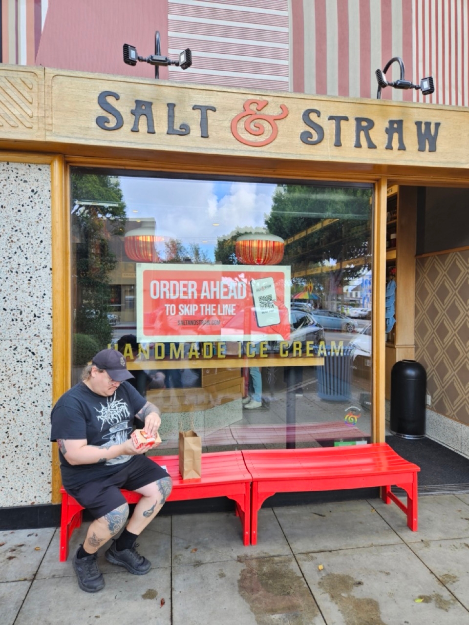Person sitting on a bench outside Salt & Straw, enjoying ice cream with a displayed sign promoting ordering ahead to skip the line