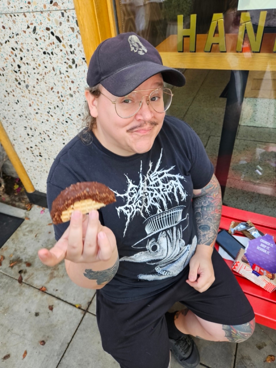 Person with tattoos, wearing a cap and glasses, smiles while holding a chocolate-covered dessert outside a shop