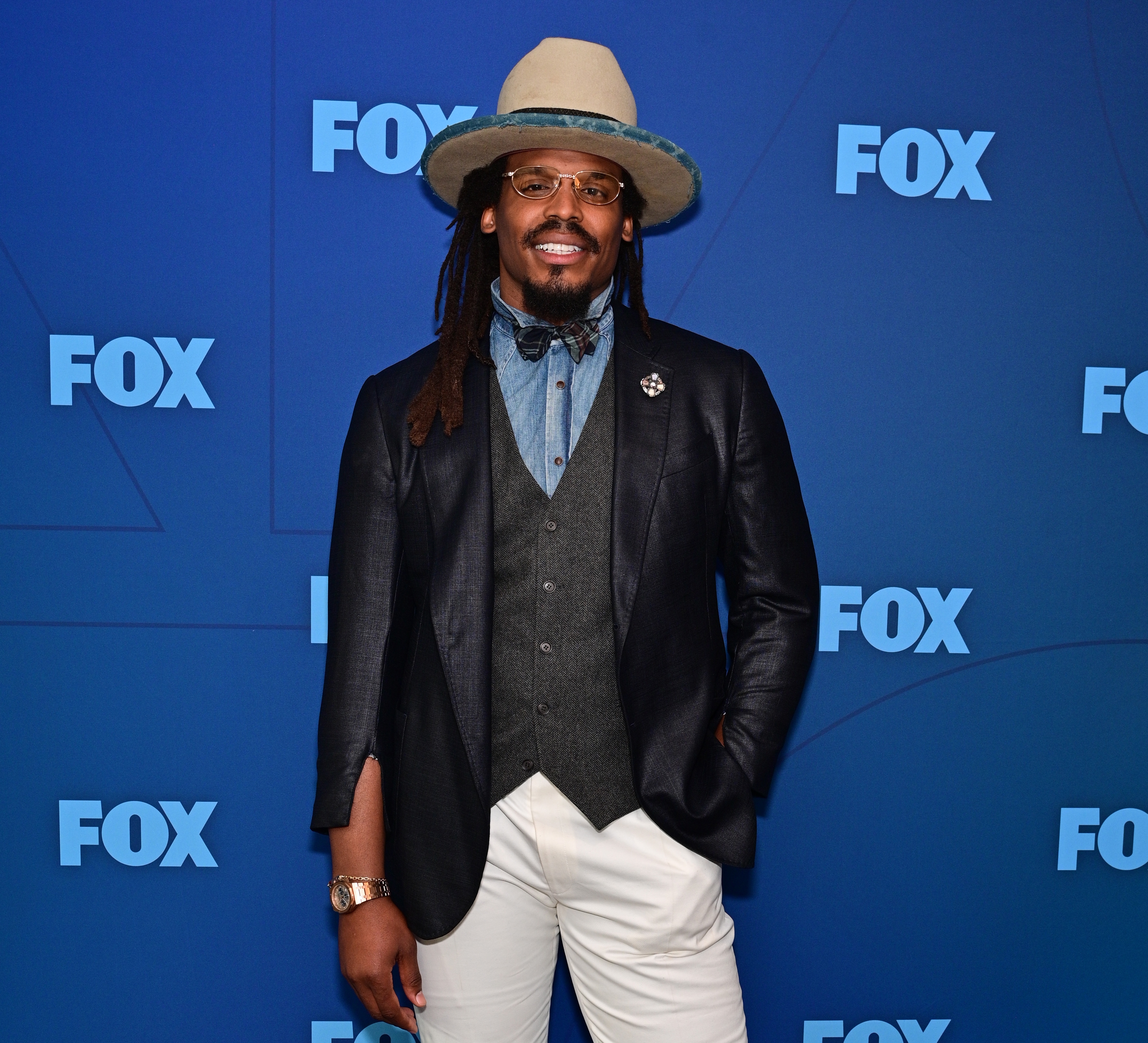 Person successful  unsocial   benignant   with wide-brim hat, acheronian  blazer, vest, bow tie, and achromatic  pants astatine  a FOX lawsuit   backdrop
