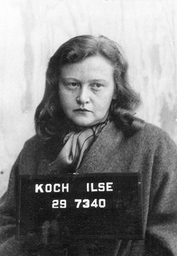 A historical mugshot of a woman wearing a coat and scarf, with a serious expression, holding a sign with her name and numbers