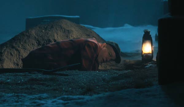 Person wearing a cap and red checkered jacket lies on the ground beside a lit lantern, suggesting a dramatic or suspenseful scene