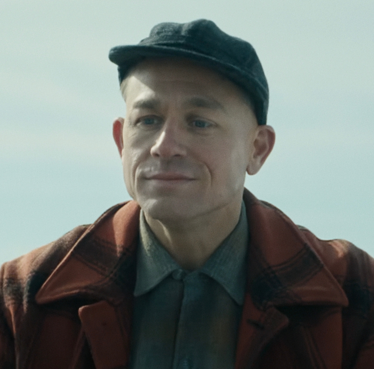 A person in a plaid jacket and cap is outdoors, smiling slightly, in a still likely from a TV or movie scene