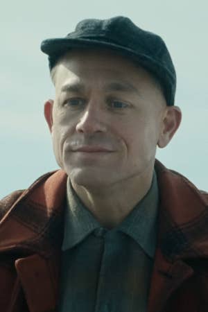 A person in a plaid jacket and cap is outdoors, smiling slightly, in a still likely from a TV or movie scene