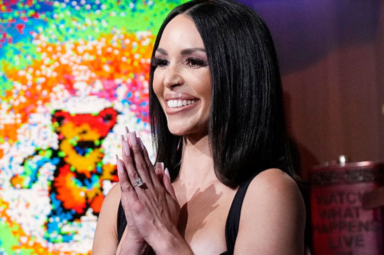 Person smiling, with straight hair, wearing a black sleeveless dress, hands clasped. Background features colorful pixel art with a bear