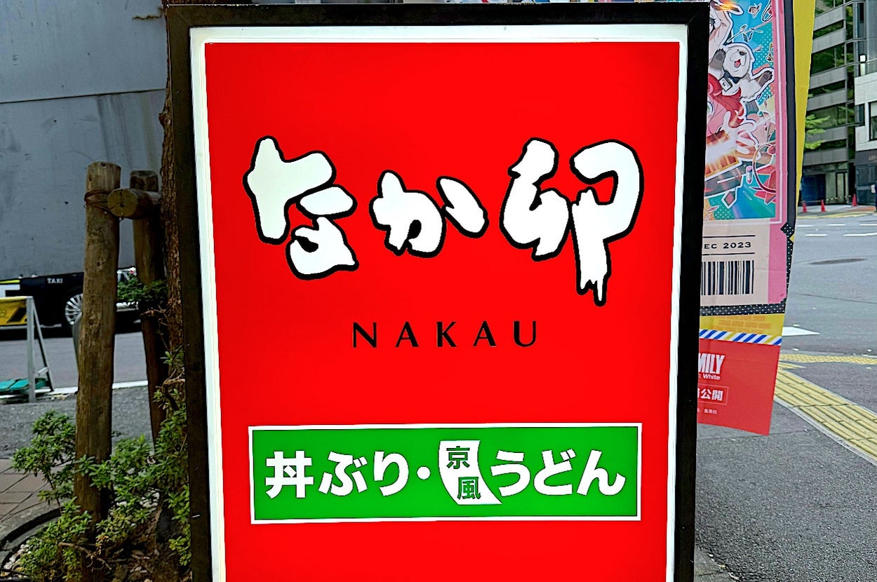 A street sign for Nakau, a restaurant, with Japanese text above and below the word "Nakau." Nearby are street and building signs