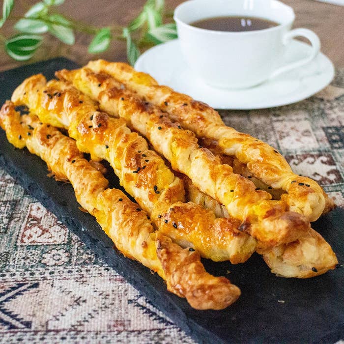 焼きたてのねじりパイのスティックが黒いプレートに並べられ、背景にコーヒーカップが置かれている。
