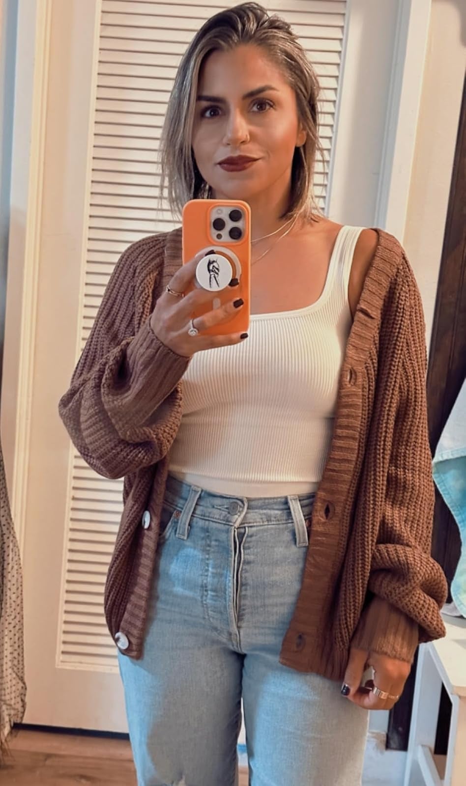 Person taking a reflector  selfie wears a cozy cardigan, achromatic  vessel  top, and jeans, holding a telephone  with a feather design